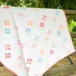 Country Crossroads Quilt Design: Simple swiss crosses sashed together on a light background. Quilt is displayed on an old farm gate outside under a large tree.