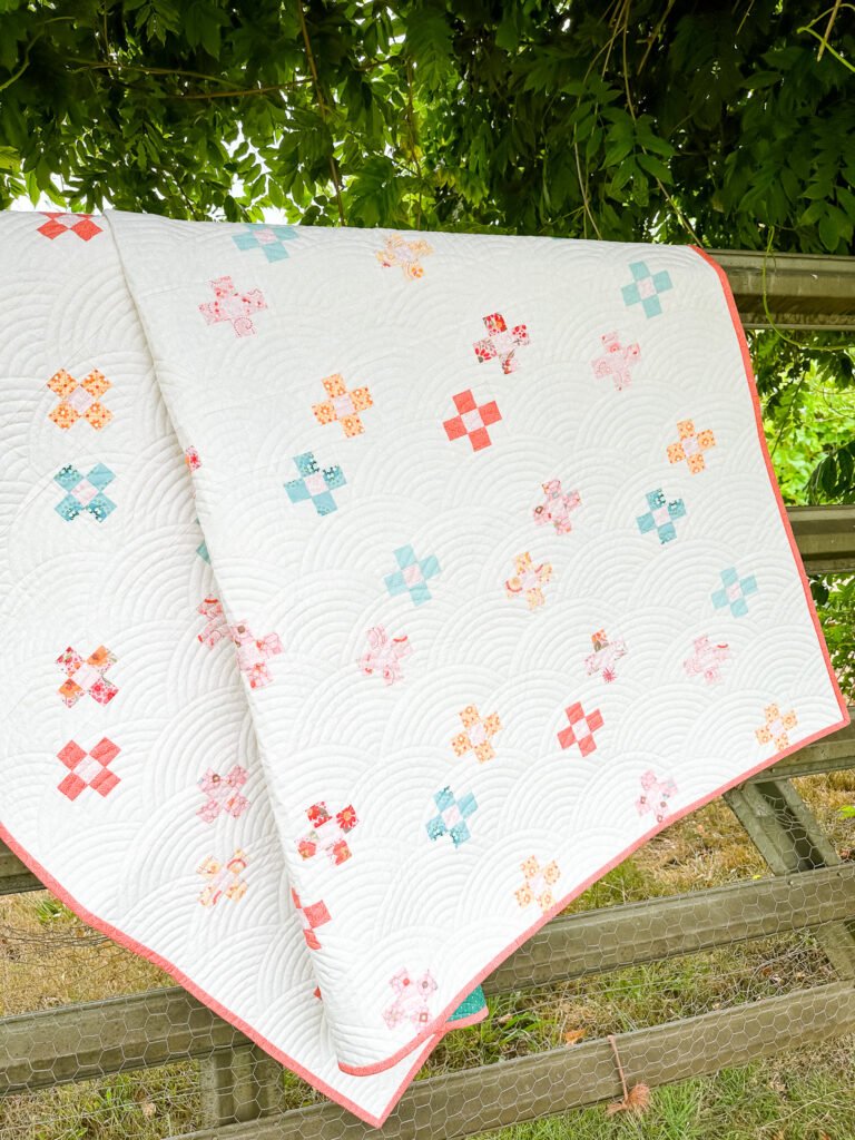 Country Crossroads Quilt Design: Simple swiss crosses sashed together on a light background. Quilt is displayed on an old farm gate outside under a large tree.